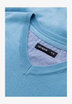 Lichtblauwe trui met een geribbelde V-hals. Beschikt over een label met de tekst "ombre" en een gladde textuur met een lichte glans.
