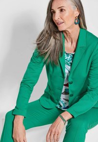 Grüner Blazer und passende Hose aus glattem Stoff, kombiniert mit einem gemusterten Oberteil mit grünen und roten Akzenten. Runde Ohrringe und ein Armband.