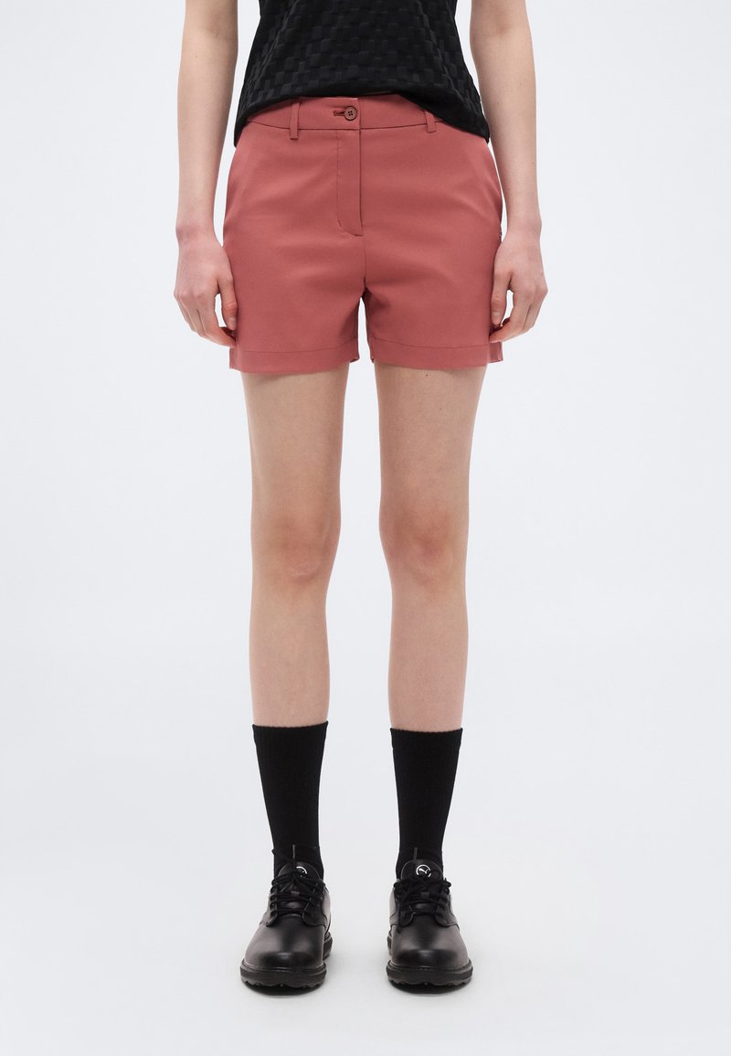 Γυναίκα που φοράει σκουρόχρωμα ρουστίκ σορτς, μαύρες ψηλές κάλτσες, μαύρα δερμάτινα παπούτσια με κορδόνια και μαύρο υφασμάτινο τοπ που έχει μπει μέσα στο παντελόνι, στέκεται σε λευκό φόντο.