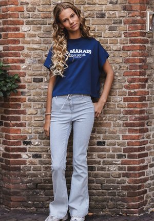 Young woman with long curly hair wearing a navy graphic t-shirt, striped flared pants, and sneakers, standing against a brick wall.