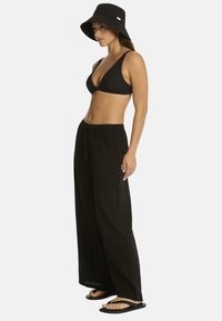 Mujer que lleva un sombrero tipo bucket negro, bralette negro, pantalones negros sueltos y chanclas negras, de pie frente a un fondo blanco liso.
