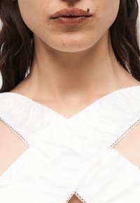 Κοντινό πλάνο στο κάτω μέρος του προσώπου μιας γυναίκας με γυαλιστερά χείλη, μια εμφανή ελιά πάνω από το χείλος και ένα λευκό τοπ με ανάγλυφα διασταυρούμενα σχέδια που φαίνεται.