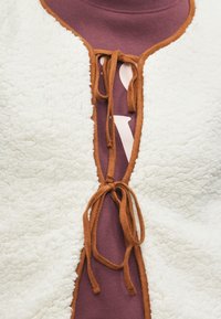 Witte fleece cardigan met bruine afwerking en strikken, over een maroon shirt met een witte grafische print. Zachte textuur en casual ontwerp elementen.