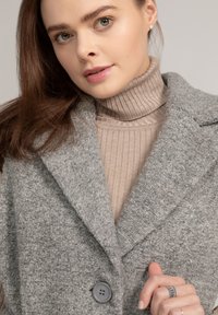 Grau strukturierter Mantel mit Reverskragen und zwei vorderen Knöpfen. Darunter ein rippierter beigefarbener Rollkragenpullover. Glatte Haut und lange braune Haare.
