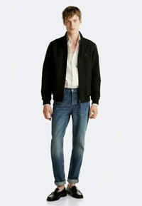 Svart bomberjakke over en hvit skjorte, parret med blå jeans. Modellen har på seg svarte loafers; jeansene er oppbrettet ved anklene.