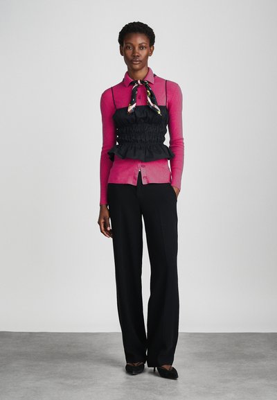 Camisa rosa de mangas longas ajustada, sobreposta a uma blusa preta de peplum com babados, combinada com calças largas pretas e sapatos pretos de bico fino.