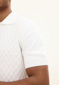 Vit stickad polo med en texturerad diamantmönster, korta ärmar och klassisk krage. Slät tyg med en figurnära design.