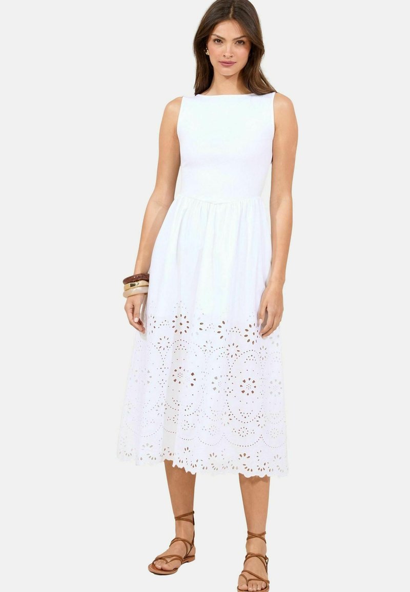 Weißes ärmelloses Kleid mit tailliertem Oberteil und ausgestelltem Rock. Der untere Teil weist laser-cut Blumenmuster auf. Ein Paar braune Sandalen mit Riemchen.