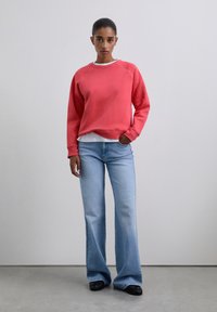Sweat-shirt corail à col rond et manches raglan, associé à un jean évasé bleu clair. Tissu décontracté, coupe confortable, et chaussures plates noires.