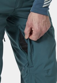 Tirkizne vodootporne hlače s mrežastim podstavljenim ventilacijskim džepovima sa patentnim zatvaračem. Kratki prikaz ruke koja povlači patent zatvarač džepa. Glatka, lagana tekstura tkanine.