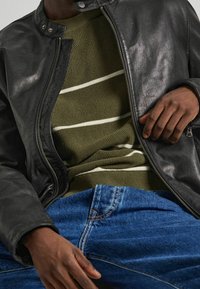 Giacca di pelle nera con colletto alto, maglione a coste verde oliva con strisce bianche sotto, e jeans in denim blu.