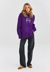 Fialová oversized mikina s přední kapsou a bílými šňůrkami, zkombinovaná s tmavými širokými džíny a hnědými pantofli.