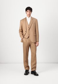 Tan three-piece suit with a single-breasted jacket, matching trousers, and a vest. Black shoes. Smooth texture, tailored fit, minimal detailing.