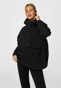 Veste noire surdimensionnée imperméable avec un col montant, une fermeture à boutons sur le devant et une grande poche ; design minimaliste avec un détail de logo subtil.