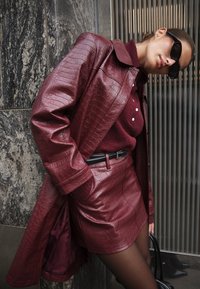 Cappotto in finta pelle bordeaux e gonna coordinata con finitura testurizzata. La cintura nera accentua il punto vita. Il modello indossa un maglione bordeaux e occhiali da sole.