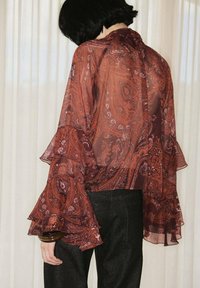 Doorzichtige blouse met paisleypatroon in rijke roest en bordeaux, met gerimpelde mouwen en een hoge hals, gecombineerd met donkere broeken.