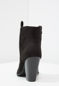 Czarna zamszowa botka na kostkę z wysokim, masywnym obcasem typu słupek i tylną pętlą do zakładania, widoczna od tyłu na białym tle.