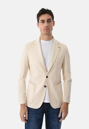 Uomo che indossa un blazer beige sopra una t-shirt bianca e jeans blu, in piedi davanti a uno sfondo bianco uniforme.