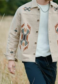 Beige jas met kleurrijke geometrische borduursels in bruin, zwart en groen. Voorzien van een knoopsluiting aan de voorkant en een klassiek kraagontwerp.