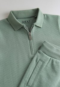 Green polo shirt with a zip collar, textured fabric, and ribbed cuff. Features a subtle patterned surface and a side pocket. Brand label visible.