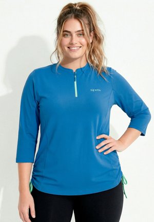 Smilende kvinde med blond hår i hestehale har en blå 3/4-ærmet atletisk trøje med lynlås og sorte bukser på, stående med hånden på hoften.