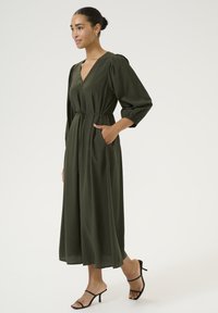 Vestido verde oliva con escote en V, mangas abullonadas, cintura con cinturón y bolsillos laterales. Tejido fluido con un corte largo y suelto, combinado con tacones negros.