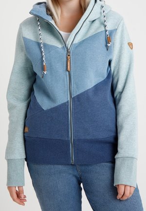 Kvinde iført en farveblokket hættetrøje med lynlås i forskellige blå nuancer med snore og tommelfingerhuller i ærmerne, kombineret med blå jeans.