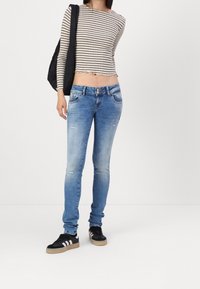 Jeans ajustados de color azul con detalles desgastados, combinados con una blusa de manga larga a rayas recortada y un bolso negro de hombro. Zapatillas negras con acentos blancos.