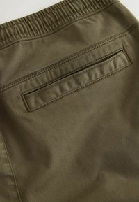 Olivgrüner Stoff mit einer glatten Textur, versehen mit einem elastischen Taillenbund und einer Gesäßtasche mit horizontaler Öffnung und Nähdetails.