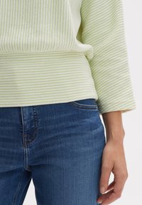 Maglione a righe verde chiaro con una consistenza morbida, caratterizzato da una vestibilità rilassata, abbinato a jeans in denim blu scuro. Una mano è appoggiata visibilmente sui jeans.