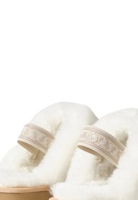 Een paar beige slippers met een witte donzige voering en decoratieve geborduurde banden op een eenvoudige witte achtergrond.