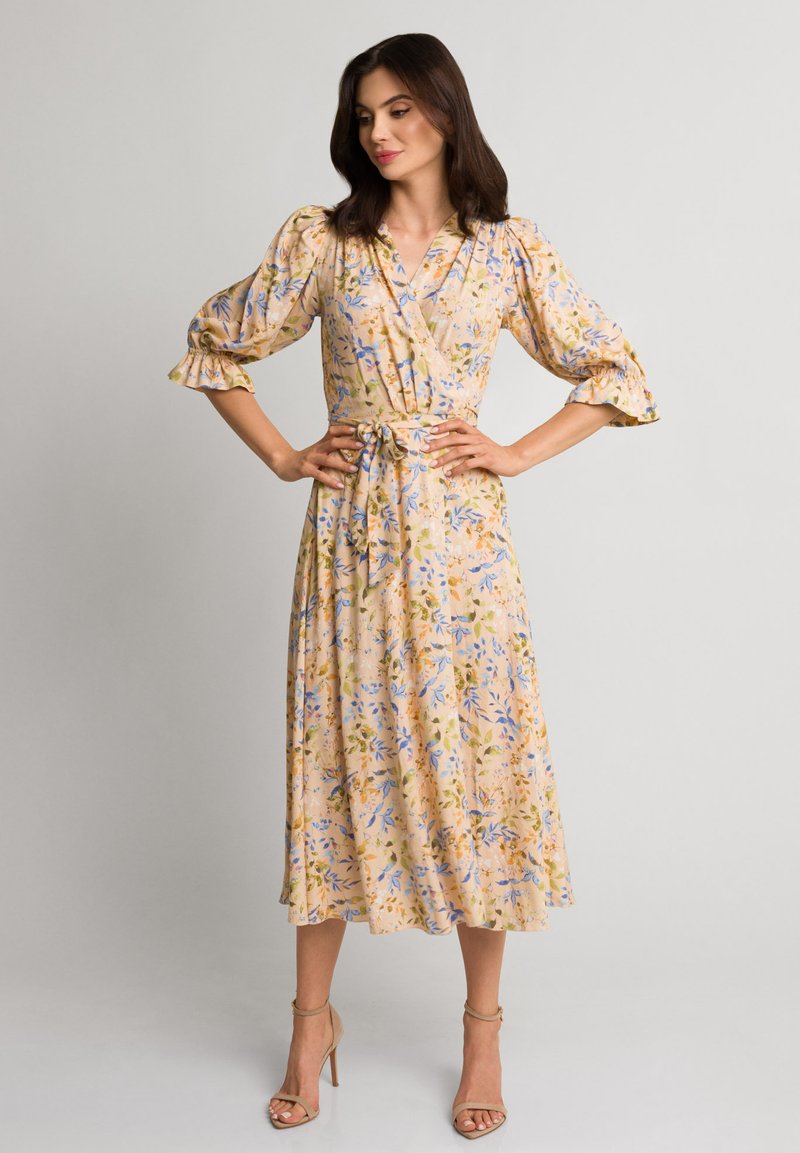 Kobieta stojąca z rękami na biodrach, ubrana w kwiatową sukienkę midi z dekoltem w serek i bufkami, oraz beżowe sandały na obcasie na jednolitym tle.