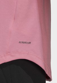 Světle růžové atletické tričko s žebrovanou strukturou a zaobleným lemem. Na bočním švu poblíž spodního okraje je černé logo "AEROREADY".