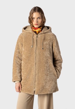 Frau trägt eine beige Kapuzen-Fleecejacke mit Händen in den Taschen und einem bunten Schal um den Hals und steht vor einem grauen Hintergrund.