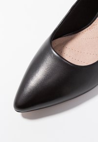 Chaussure plate en cuir noir à bout pointu avec un extérieur lisse, des côtés ajourés et une semelle intérieure beige rembourrée présentant de petites perforations.