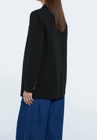 Černý na míru šitý blejzr s klopami, který má jediný zadní rozvent a manžety na knoflíky, kombinovaný s volnými modrými džínovými kalhotami.