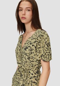 Kurzärmliges, V-Ausschnitt-Kleid in Olivgrün mit einem schwarzen Blumenmuster, an der Taille mit einem passenden Band gerafft. Glatte Textur.