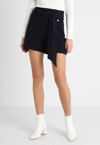 Shorts bleu marine taille haute avec détail ceinturé, présentant une texture lisse et une coupe ajustée. Associés à des bottines blanches et un haut moulant.
