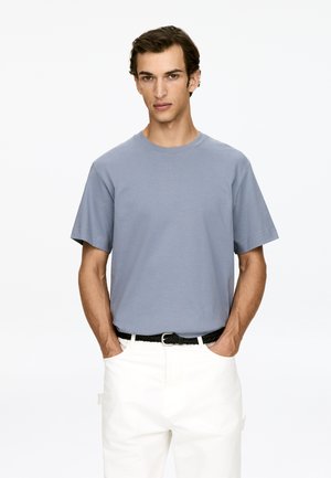 T-shirt en coton bleu clair à manches courtes et col rond, associé à un pantalon blanc et une ceinture tissée noire, les mains dans les poches.