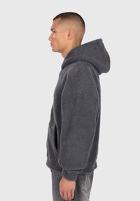 Mand iført en mørkegrå fleece hættetrøje med forlommer, som står i profil mod en ensfarvet lys grå baggrund.