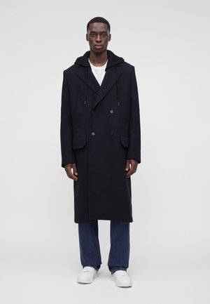 Homme portant un long manteau noir croisé avec capuche, jean bleu, baskets blanches et chemise blanche, debout devant un fond uni.
