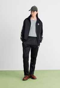 Fato de treino preto com detalhes cinzas. Casaco com fecho éclair e logo, calças a condizer e sapatos castanhos. Camiseta cinza por baixo. Boné usado.