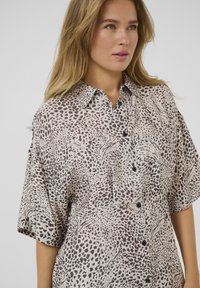 Bluza kratkih rukava, na kopčanje, od bež tkanine sa tamnim leopard uzorcima i apstraktnim motivima. Ima klasičan ovratnik i crne dugmadi.