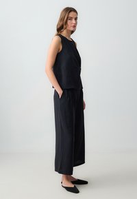 Débardeur noir sans manches avec un design à l'avant torsadé, associé à un pantalon noir oversized et fluide. Le tissu semble doux et lisse.