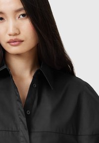 Camicia nera con bottoni, caratterizzata da un colletto strutturato, nota per la sua superficie liscia e il fit rilassato. Presenta una leggera lucentezza e bottoni nascosti.