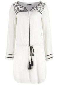 Robe blanche à manches longues avec broderie noire sur le yoke et les poignets, bordure noire verticale, et lien à pompons noirs à la taille.