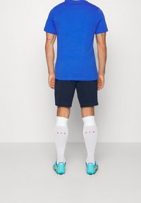 Camiseta de algodón azul, pantalones cortos azul marino, calcetas blancas hasta la rodilla con texto rosa y botas de fútbol turquesa, vistas desde la espalda.