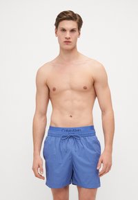 Mėlyni plaukimo šortai, pagaminti iš lengvos medžiagos, elastinga juosmens juosta su raišteliu ir Calvin Klein logotipas ant juosmens juostos. Lygus paviršius, kasdienis dizainas.