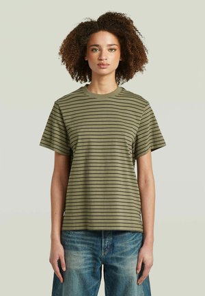 Jonge vrouw met krullend haar draagt een groen-zwart gestreept t-shirt en blauwe jeans, staand tegen een effen lichte achtergrond.