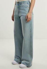 Jeans de culoare albastră deschisă, cu talie înaltă și picioare largi, cu un finisaj uzat, având buzunare frontale și detalii de cusătură, asortați cu adidași albi.